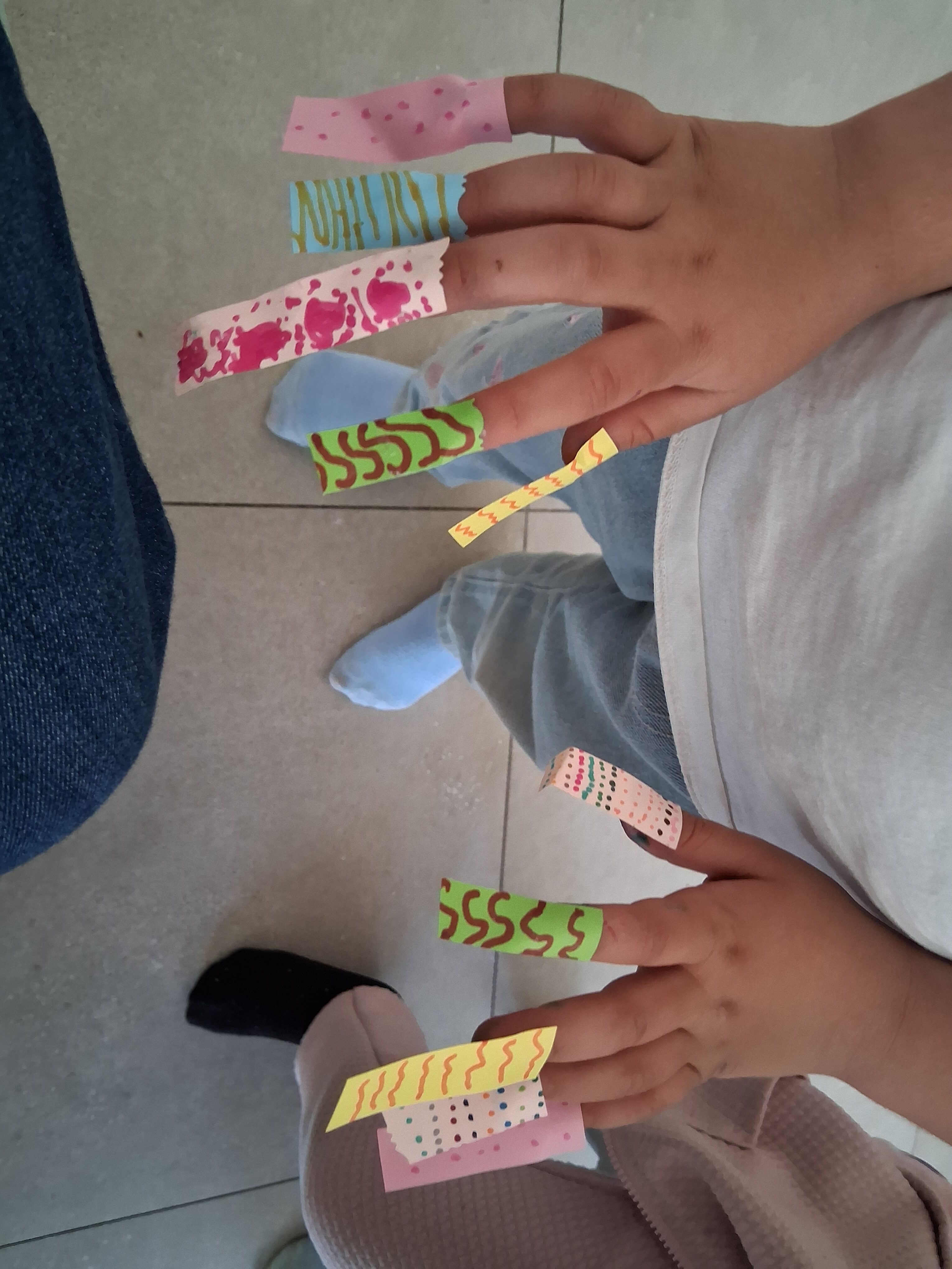 Twee kinderhanden met gekleurde papierrandjes rond de vingertoppen, staand op een betegelde vloer.