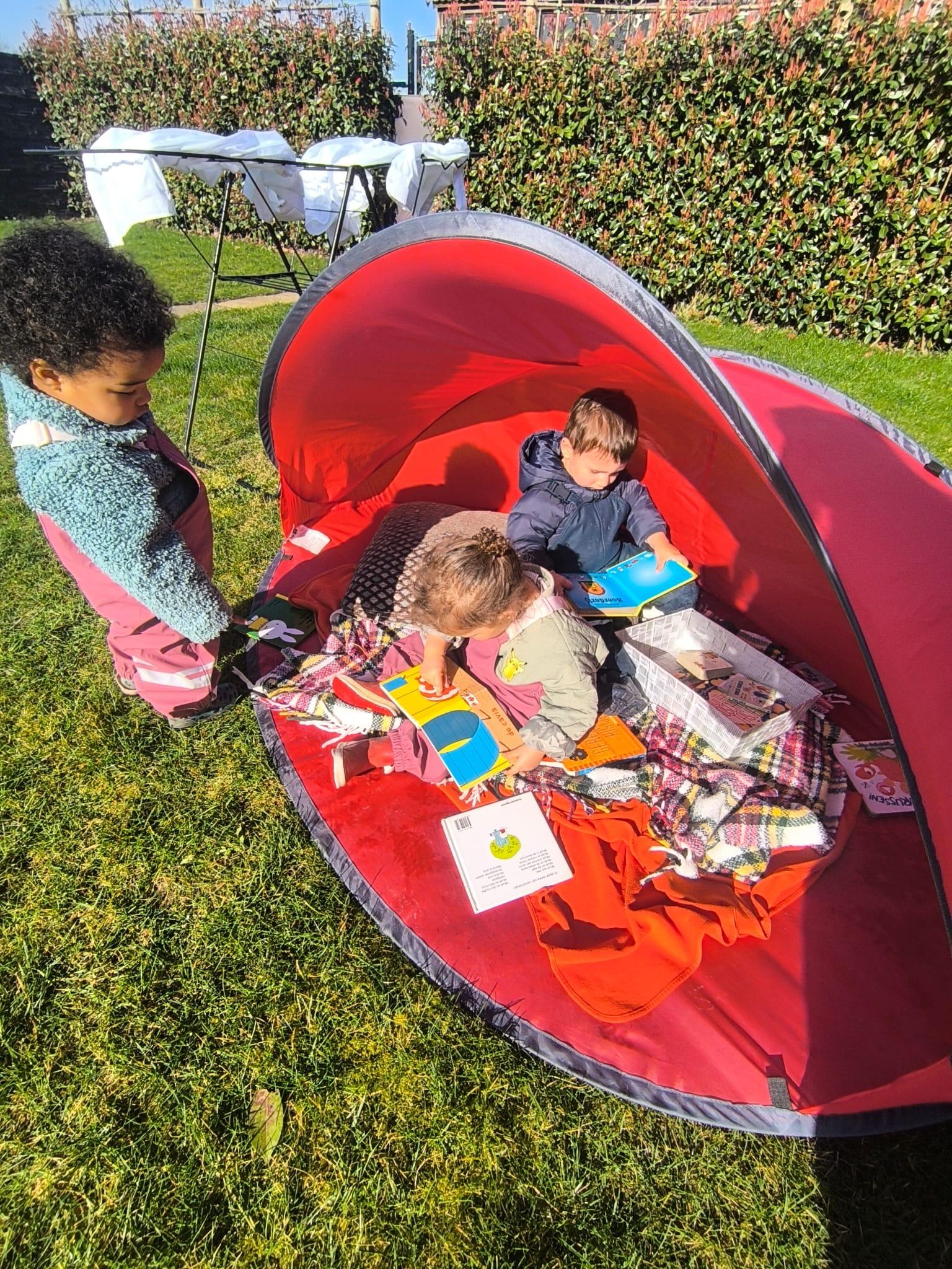 Drie kinderen spelen buiten in een rode tent met boeken en dekens op het gras.